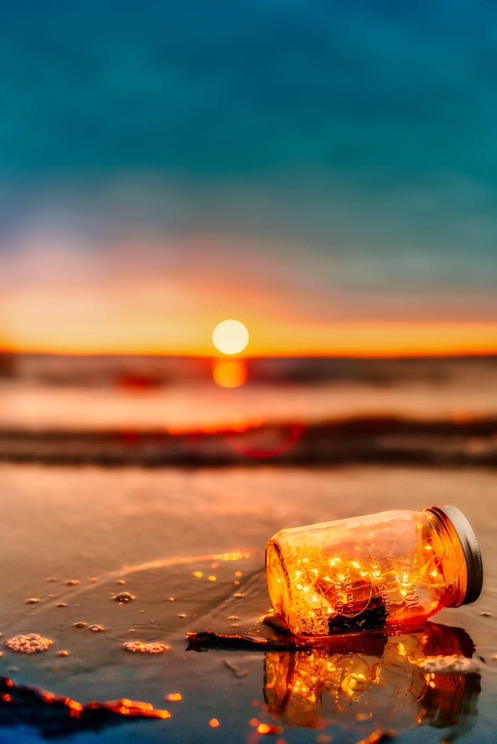 A lit jar with lights sits on a beach, creating a tranquil scene during sunset.